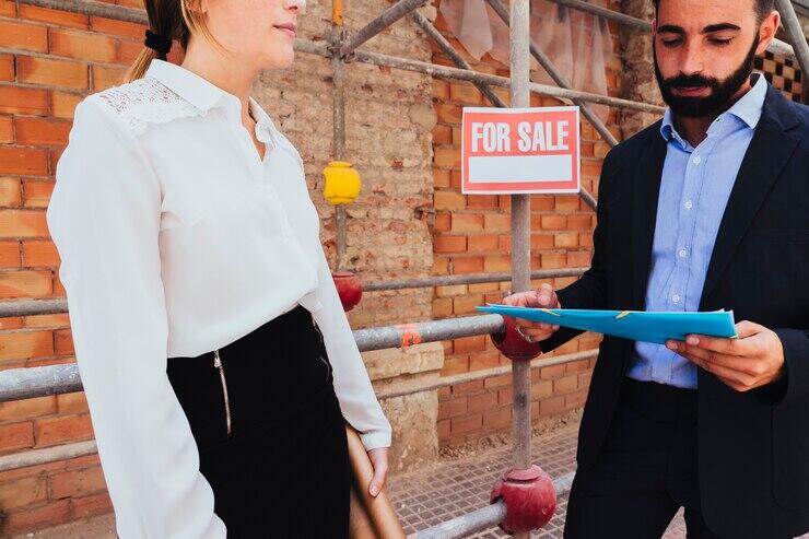 Businesswoman and real estate agent reviewing documents at a construction site. "For Sale" sign visible.